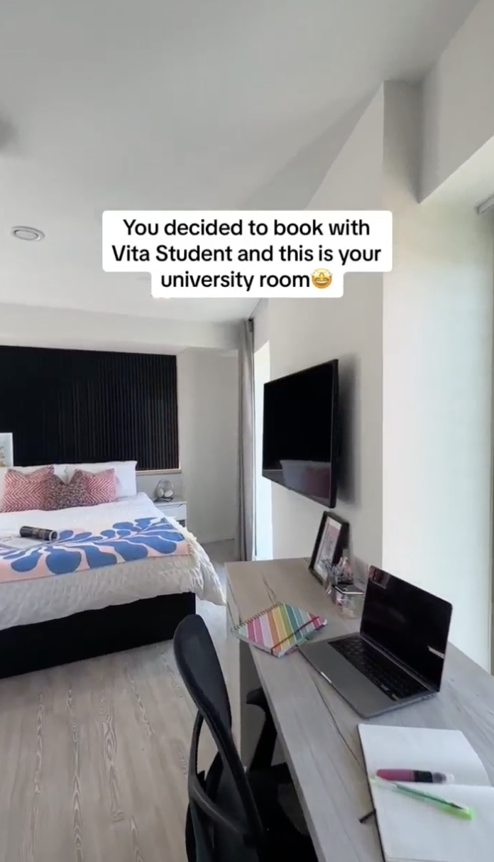 Modern dorm room with large bed, geometric patterned bedding, wall-mounted TV, and study desk. The desk holds a laptop, stationery, and a photo frame. Large window on the right lets in ample natural light. A caption reads: "You decided to book with Vita Student and this is your university room.